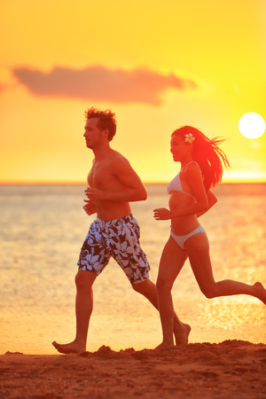 Jogging couple running exercising at sunset beach. Fitness people, man and woman training outside by the ocean sea in beautiful sunset or sunrise in full body length in summer.の写真素材