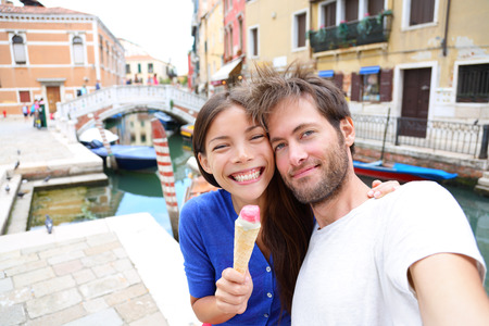 Couple in Venice, eating Ice cream taking selfie self-portrait photo on vacation travel in Italy. Smiling happy Asian woman and Caucasian man in love having fun eating italian gelato food outdoors.の写真素材