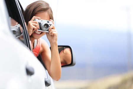 Woman tourist taking photo in car with camera driving on road trip travel vacation. Girl passenger taking picture out of window with vintage retro camera.の写真素材