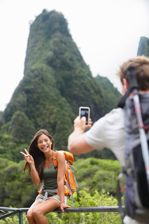 Couple taking photos having fun lifestyle hiking on Hawaii in outdoor activity. Woman and man hiker taking photo pictures with smart phone camera. Iao Valley State Park, Wailuku, Maui, USA.の写真素材