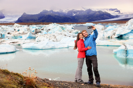 Iceland - couple taking selfie self portrait photo by Jokulsarlon glacial lagoon / glacier lake. Happy tourists on travel enjoying beautiful Icelandic nature landscape with Vatnajokull in backround.の写真素材