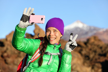 Woman hiker taking selfie photo using smartphone while hiking in winter jacket and clothing enjoying outdoor activity. Woman hiker taking self-portrait picture with smart phone camera.の写真素材