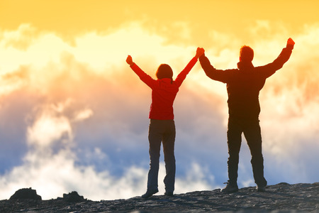 Happy winners reaching life goal - success people at summit. Business achievement concept. Two person couple together arms up in the air of happiness with accomplishment in the clouds at sunset.の写真素材