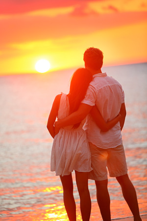 Honeymoon couple romantic in love at beach sunset. Newlywed happy young couple hugging enjoying ocean sunset during travel holidays vacation getaway. Interracial couple, Asian woman, Caucasian man.の写真素材
