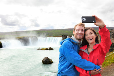 Selfie couple taking smartphone picture of waterfall Godafoss outdoors on Iceland. Couple visiting famous tourist attractions and landmarks in Icelandic nature landscape. Mixed race couple having fun.の写真素材