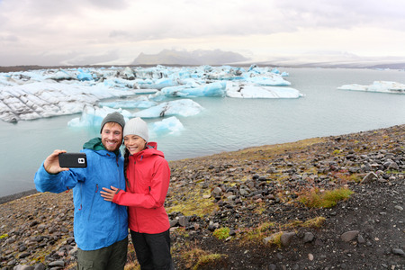 Travel couple taking selfie self portrait on Iceland photo by Jokulsarlon glacial lagoon / glacier lake. Happy tourists enjoying beautiful Icelandic nature landscape with Vatnajokull in background.の写真素材