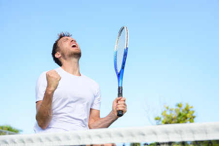 Tennis player man winning cheering celebrating victory. Winner man happy in celebration of success and win. Fit male athlete on tennis court outdoors holding tennis racket in triumph by the net.の写真素材