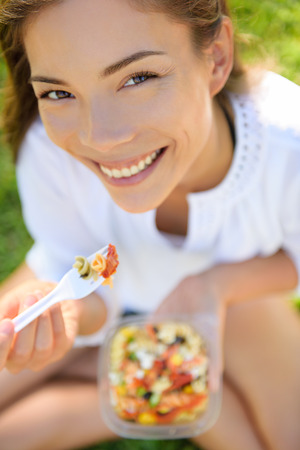 Woman eating gluten free pasta salad. Portrait of girl eating lunch takeaway in park outdoors. Biracial Asian Caucasian female model in her twenties smiling happy looking at camera.の写真素材