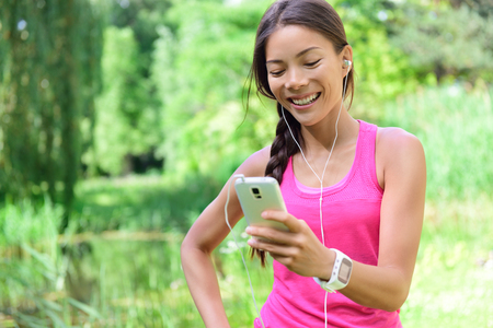 Woman runner sharing running data on social media after exercise. Girl listening to music on smart phone after jogging run in city park. Female jogger with earphones, smartphone and heart rate monitor.の写真素材