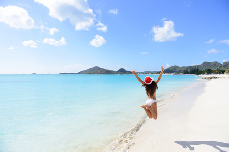 Christmas beach travel vacation woman in Santa hat jumping of joy on tropical Caribbean winter holidays getaway. Beautiful girl in bikini having fun under the sun traveling.の写真素材