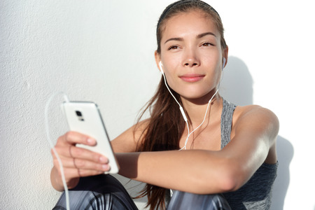 Young woman listening to music with earphones on smart phone app for fitness motivation. Athlete runner in sportswear relaxing sitting getting inspired. Asian mixed race model.の写真素材