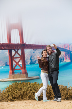 Tourists couple taking selfie photo in San Francisco by Golden Gate Bridge. Interracial young modern couple using smart phone by famous amaerican landmark. Asian woman, Caucasian man.の写真素材