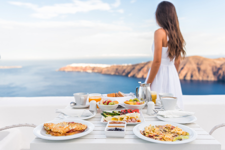 Breakfast table and luxury travel woman on santorini. Well balanced perfect breakfast table served at resort. Female tourist is looking at beautiful view of sea and caldera enjoying her vacation.の写真素材