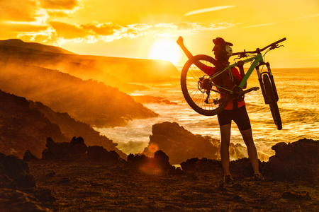 Success, achievement, accomplishment and winning concept with cyclist mountain biking. Happy MTB woman cycling raising arms lifting bike by sea during sunset cheering and celebrating at summit top.の写真素材