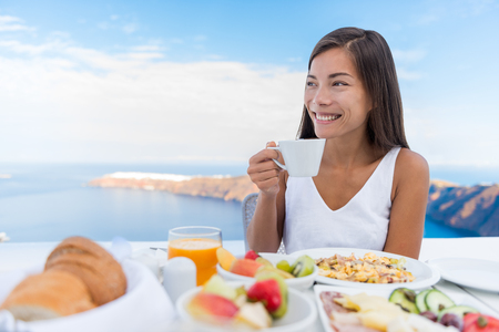 Asian Woman drinking hot drink at morning Breakfast table. Beautiful elegant lady having coffee cup on luxury hotel terrace with sea view at resort restaurant on Oia Santorini island, Greece.の写真素材