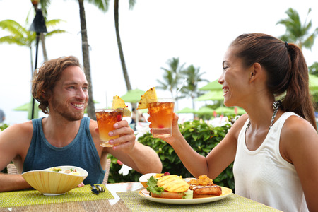 Happy multiracial couple toasting cheers with alcoholic hawaiian drink, Mai Tai, Hawaii experience. Summer travel holidays, people enjoying local food meal at outdoor terrace restaurant of resort.の写真素材