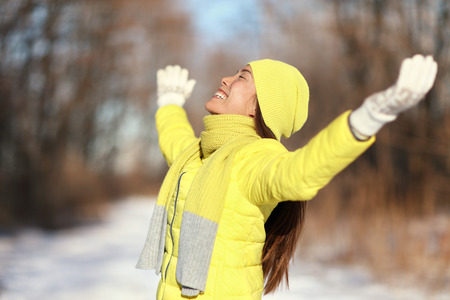 Happy snow freedom carefree woman. Relaxing Asian woman soaking up the sun on a sunny winter day with arms up in forest nature outdoors in yellow down jacket with knit hat and warm scarf.の写真素材