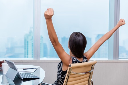 Winning office worker working woman with arms raised up outstretched in success of career goal achievement. Happy successful businesswoman looking at city view in work window.の写真素材
