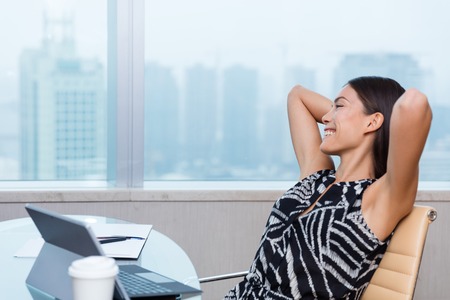 Work satisfaction office woman relaxing at desk. Happy Asian businesswoman worker of goal success or enjoying her job achievement looking at skyline. Working from home freedom concept.の写真素材