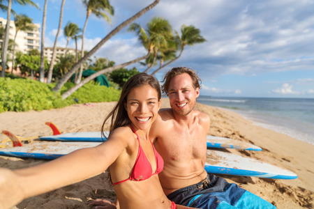 Happy young interracial couple taking phone selfie picture relaxing sitting near surfboards on beach holiday having fun doing surfing activity class. Travel vacation destination watersports.の写真素材