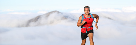 Trail runner man athlete running in mountains outdoor nature with mountain peak in clouds in background. Panorama horizontal banner landscape crop for copyspace.の写真素材