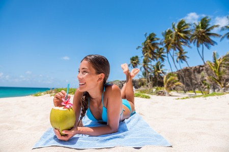 Happy girl lying down sun tanning on beach vacation. Asian woman relaxing sunbathing drinking fresh coconut water. Tropical travel destination, Suntan in summer holiday.の写真素材