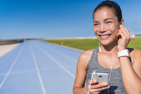 Asian runner woman listening to running motivation music with earbuds on her mobile phone app for run on track and field blue lanes tracks in athletics stadium. Athlete enjoying fitness active life.の写真素材