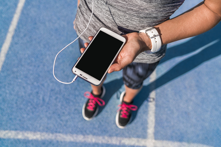 Runner girl holding smartphone using touchscreen for choosing music or texting sms on app before running on track. Female athlete woman feet and leg closeup with smartwatch and hand touching display.の写真素材