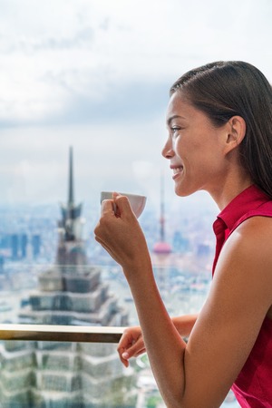 Asian woman enjoying afternoon high tea in luxury cafe or fancy restaurant with city view of Shanghai's landmark skyscrapers in Pudong, China. Chinese tourist lady drinking hot cup of coffee relaxing.の写真素材