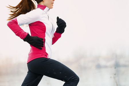 Winter running athlete woman on cold run jogging fast with speed and sprint on outside workout wearing warm clothing gloves, winter tights and wind jacket in snow weather.の写真素材