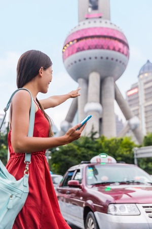Asian woman hailing a Shanghai taxi cab using mobile phone app. Lady tourist or businesswoman with hand up calling a car in from of famous China landmark using smartphone to request a ride online.の写真素材