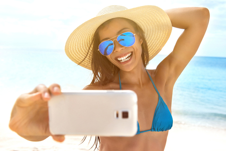 Selfie summer beach vacation travel girl taking self-portrait photo with phone on sun tan holiday. Woman wearing beach hat, mrror sunglasses, blue swimsuit.の写真素材