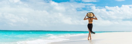 Luxury travel summer beach vacation woman walking in black beachwear skirt and hat on paradise white sand Caribbean beach. Lady tourist on Caribbean holiday vacation resort. Banner panorama landscape.の写真素材