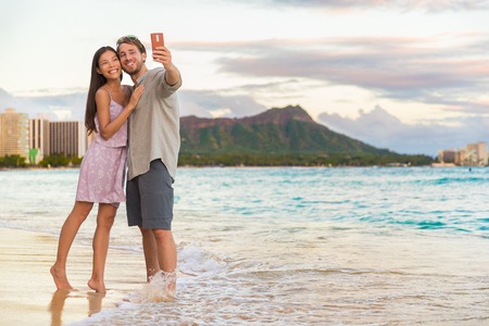 Couple walking on beach at sunset taking selfie picture on mobile phone relaxing together on Waikiki beach, Honolulu, Hawaii travel vacation. Romantic holiday destination for honeymoon.の写真素材