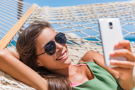 Vacation woman using smart phone taking smartphone selfie. Girl relaxing on beach hammock sun tanning in sunglasses smiling using cellphone to take pictures and text sms messages.の写真素材