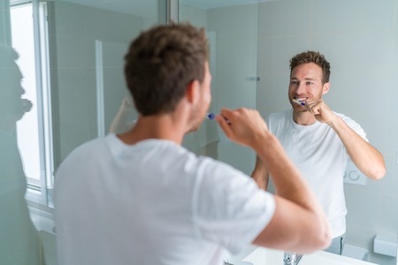 Man getting ready in the morning doing hygiene routine brushing his teeth looking in mirror of home bathroom using toothbrush in for clean dental oral care.の写真素材