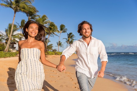 Happy newlyweds couple running on the beach holding hands, honeymoon travel. Asian caucasian multiracial people having fun together.の写真素材