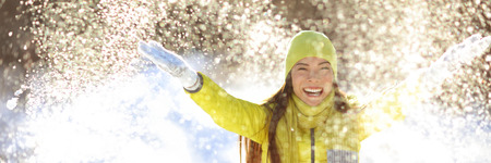 Happy winter fun woman throwing snow banner. Panorama crop of outdoor lifestyle girl playing in snow outside laughing in yellow coat, hat, gloves and scarf.の写真素材