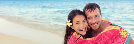 Happy couple summer fun hugging under towel on beach vacation. Laughing young people cuddling in Hawaii holiday banner panorama. Asian girl, Caucasian man interracial friends.の写真素材