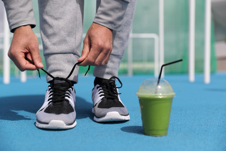 Gym fitness man getting ready tying running shoes with green smoothie detox beverage. Closeup of running trainers with plastic cup of breakfast vegetable juice. Sport active lifestyle.の写真素材