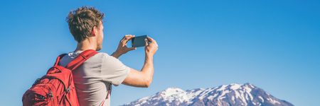 Travel tourist hiking man taking picture with phone of mountains landscape on summer hike adventure. Panorama banner with copy space on blue sky background.の写真素材