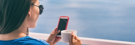 Phone woman drinking coffee cup on Europe summer vacation relaxing girl at cafe reading cellphone at outdoor terrace view of Ocean in Santorini, Greece. European destination holiday. Banner panorama.の写真素材