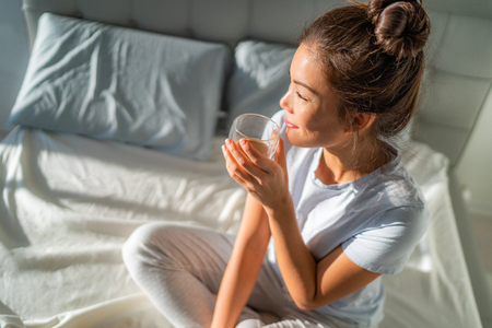 Morning breakfast in bed happy Asian woman drinking hot coffee mug relaxing sitting on mattress. Weekend relaxation wellness.の写真素材
