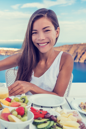 Woman eating healthy breakfast at restaurant table with Mediterranean sea view background from hotel terrace. Smiling Asian girl, vegetarian fruits and vegetables food retreat.の写真素材