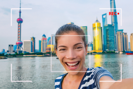 Happy China travel selfie Asian tourist woman vlogging online recording videoblog vlog video. Smiling young girl holding camera phone at the Bund at Shanghai's skyline of skycrapers.の写真素材