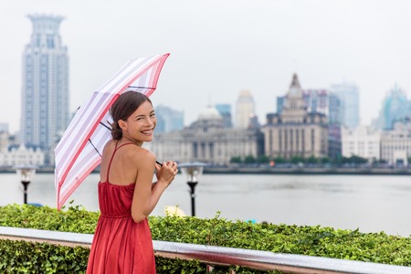 Beautiful young chinese woman enjoying walk on Pudong waterfront overlooking Huangpu district over the Bund, joyful under umbrella on a rainy summer day. Tourist travelling in Shanghai, China.の写真素材