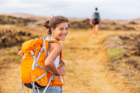 Happy young Asian hiker girl hiking with friend in mountain nature trail wearing orange backpack smiling looking back enjoying travel holiday in summer. Adventure wanderlust lifestyle. Healthy woman.の写真素材