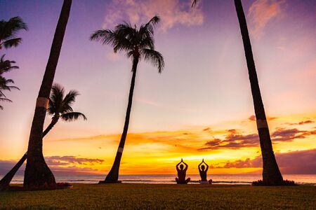 Yoga retreat on tropical summer holiday meditation people couple meditating in lotus pose with praying hands in sunset glow silhouettes on beach.の写真素材