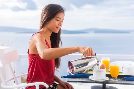 Coffee woman pouring tea in mug for breakfast at hotel room by Mediterranean sea on Europe vacation travel. Happy Asian girl enjoying morning brunch on couple honeymoon.