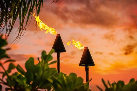 Hawaii luau party Maui fire tiki torches with open flames burning at sunset sky clouds at night. Hawaiian cultural travel vacation background.の写真素材